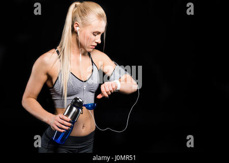 Junge blonde Sportlerin Kopfhörer halten Sie Flasche und mit Blick auf smartwatch Stockfoto