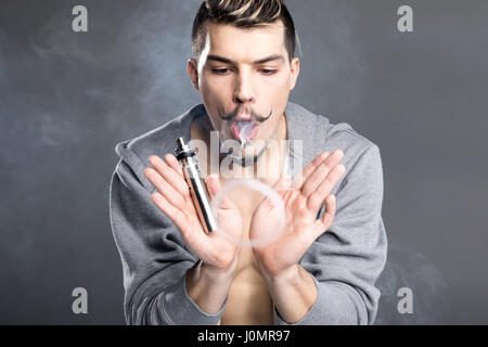 Junge bärtigen Mann, der elektronischen Zigarette und Dampfen auf grau Stockfoto