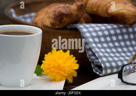 Nahaufnahme der einen Korb mit Croissants, eine Tasse Kaffee mit einem kleinen gelben Blume, ein offenes Buch und eine Brille. Stockfoto