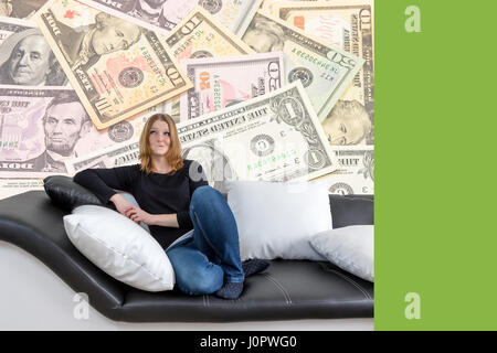 Grinsende langhaarige junge Frau sitzt auf einer schwarzen und weißen Couch mit schwarzen und weißen Kissen. Frau ist auf der Suche nach oben auf die US-Dollar-scheine ich Stockfoto