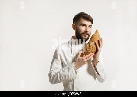 Gut aussehend lächelnd BAKER riecht ein frisch gebackenes Brot in der Küche der Bäckerei. Gut aussehend lächelnd BAKER riecht ein frisch gebackenes Brot Oon weißer zurück Stockfoto