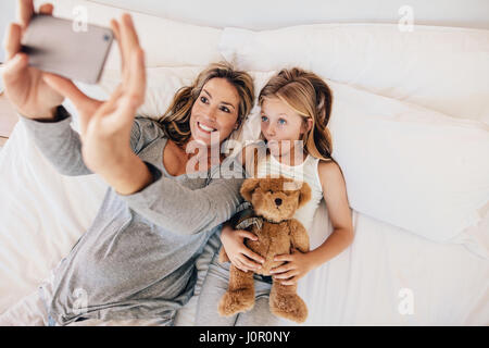 Mutter und Tochter am Bett liegen und unter Selbstbildnis mit Handy. Frau unter Selfie mit einem kleinen Mädchen halten Teddybär im Bett.