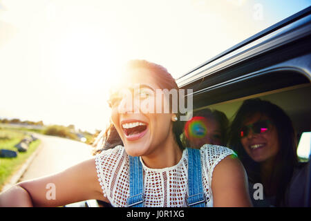 Glücklich begeistert junge Frau lehnt sich aus einem Auto Lachen Hintergrundbeleuchtung durch eine helle Fackel von der Morgensonne wie sie und ihre Freunde gehen auf eine Straße Reise Zusam Stockfoto