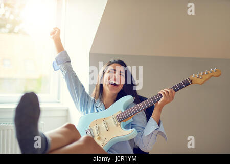 Schöne single singen Frau Gitarre spielen mit angehobenem Arm und die Füße auf den Tisch im Zimmer mit hellen Fenster Stockfoto