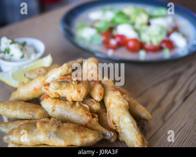 Frittierte Sardellen, Sprotten in Teig mit einer Zitronenscheibe und Bad auf einem Holztisch mit Tomaten, Feta-Käse und Avocado-Salat serviert. Stockfoto