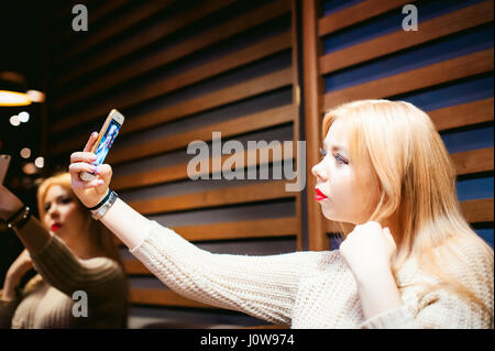 Junge, blonde Frau in einem gestrickten Pullover, ein Selfie Foto auf einem Smartphone, sitzen an einem Tisch in einem Café, Zeit allein verbringen, tun Internet Kommunikations- Stockfoto