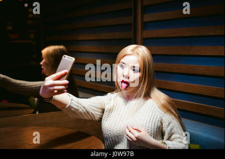 Junge, blonde Frau in einem Strickpullover, Zunge, sitzen an einem Tisch in einem Café, verbringen Zeit al zeigt, ein Selfie Foto auf einem Smartphone emotional Stockfoto