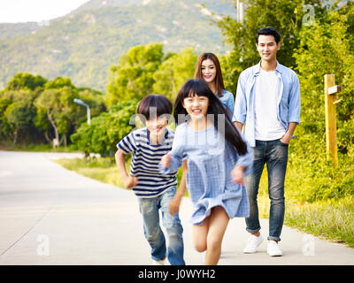 zwei asiatische Kinder laufen im Park, während ihre Eltern liebevoll beobachten. Stockfoto