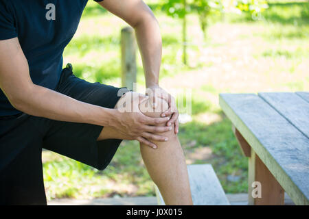 Closeup 7/8 Porträt, Mann in Schwarz Shirt und Shorts holding Knie mit schweren Schmerzen, vereinzelte Bäume und Picknickbank außerhalb Hintergrund. Stockfoto