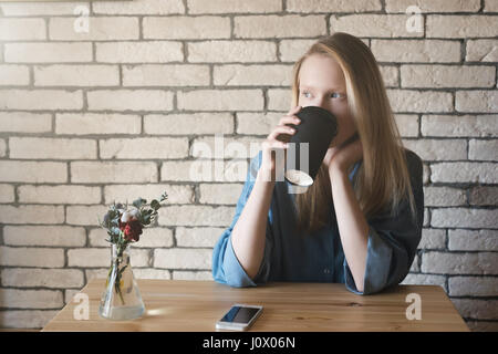 Junge Blondine im blauen Hemd sitzt am Tisch in einem Café auf dem Lügen eine Telefon auf einem gemauerten Wand Hintergrund. Eine Mädchen trinkt Kaffee aus einer Tasse schwarzem Papier ein Stockfoto