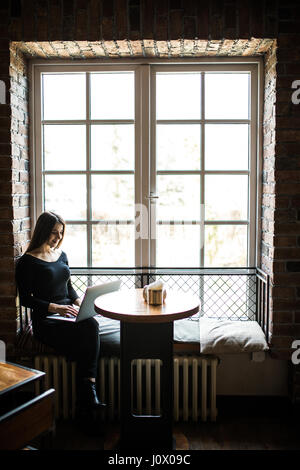 Silhouette der junge Frau sitzt am Tisch vor Fenster und nutzt Laptop. Stockfoto