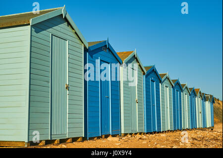 Charmouth, Dorset und eine Linie von Strandhütten in abwechselnden hellen und dunklen blauen Farben auf der Rückseite der Kiesstrand an einem Sommerabend. Stockfoto