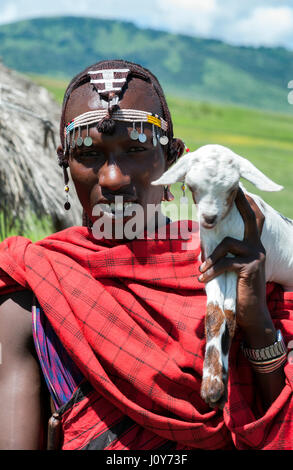 Porträt eines jungen Ostafrikas tansanische Maasai-Krieger im Stammeskopf Schmuckperlen und Silberschmuck, traditionelle rote Shuka Robe und weiße Ziege. Stockfoto