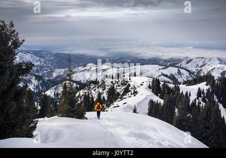 Wanderer mit Rucksack in gelben Jacke in den verschneiten Bergen mit Blick auf die Stadt zu Fuß Stockfoto