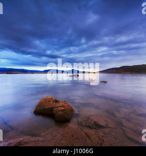 Trübe Sonnenaufgang über Jindabyne See in New South Wales schneebedeckten Berge Region Australiens. Klares Wasser des Bergsees felsigen Botton zeigt und reflektiert das Sonnenlicht. Stockfoto