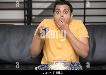 Closeup Portrait, junger Mann im gelben T-shirt, auf schwarzem Ledercouch sitzen, vor dem Fernseher, Fernbedienung, halten, überrascht, was er sieht, kaute popcor Stockfoto