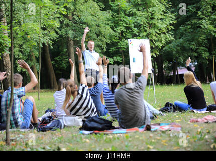 Eine Gruppe von jungen Studenten in den Park. Ansicht eines Mannes gestikulieren mit den Händen gegen einen defokussierten Gruppe von Menschen, die vor ihm saß auf dem Rasen stehen. Stockfoto