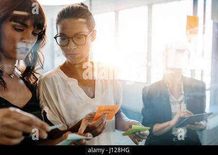 Business-Frauen diskutieren vor der Glaswand mit Post, die er nimmt zur Kenntnis und Aufkleber. Corporate-Profis mit Klebstoff Noten für ein Brainstorming in m Stockfoto