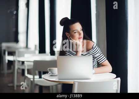 Porträt von einer attraktiven Frau in einigen Coffee-Shop mit ihrem Laptop sitzen und denken über ein Thema beim Blick durch das Fenster. Stockfoto