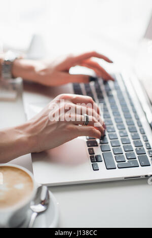 Nahaufnahme Foto von weiblichen Händen tippen auf Laptoptastatur mit einer Tasse Cappuccino gemustert. Stockfoto