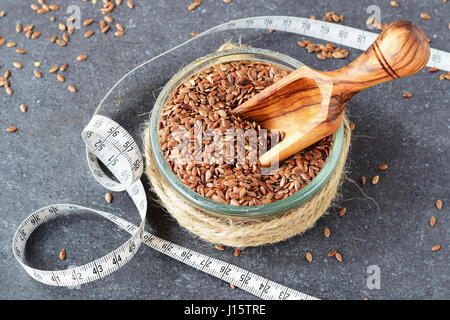 Eine Glasschüssel mit Leinsamen, Oliven Holz Kugel drin, mit Maßband abstrakt grau unterlegt. Gesunde Ernährung, Konzept, Diät, Schönheit. Stockfoto