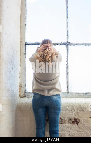 Rückansicht einer gestressten blonden Frau stehend durch die Fenster Kopf in Händen Stockfoto