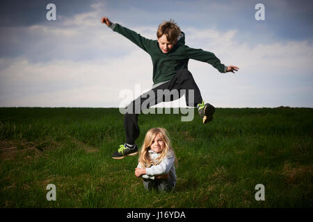 Bruder Schwester auf Wiese, bewölkten Himmelshintergrund überspringen. Kinder Sommer outdoor-Aktivitäten. Hoch und weit springen Stockfoto