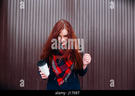 Schöne glückliche junge Frau hält einen Pappbecher und Kaffee trinken. Fröhliche Frau mit roten langen Haaren auf der Straße trinken Kaffee am Morgen Stockfoto