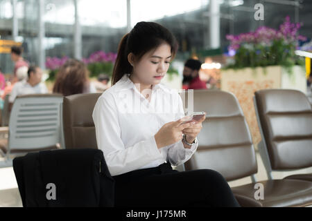 Junge asiatische Geschäftsfrau mit Smartphone beim Sitzen im Flughafen-Terminal für das Boarding warten. Stockfoto