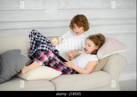 Zwei Kinder, der junge und das Mädchen liegen auf dem Sofa mit dem Tablett  in der Hand. Sie mit Interesse auf den Bildschirm schauen. Hobby der  modernen Kinder Stockfotografie - Alamy