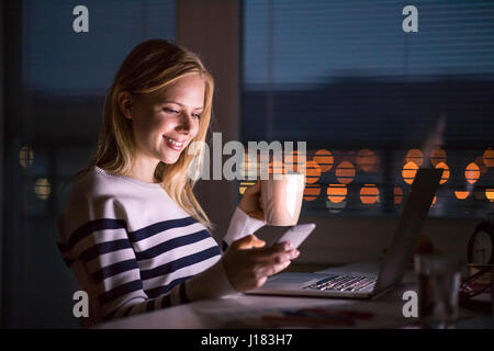 Frau an der Rezeption, mit Smartphone, nachts am Laptop arbeiten. Stockfoto