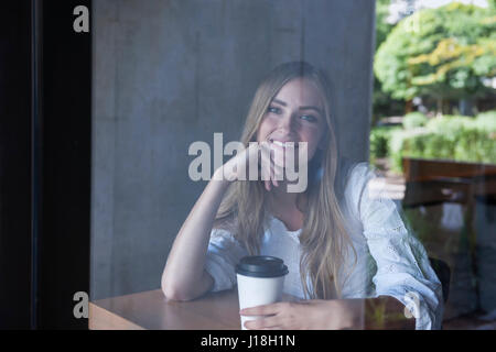 Lächelnde Mädchen sitzen im Café mit Kaffee durch Fensterglas von außen fotografiert Stockfoto