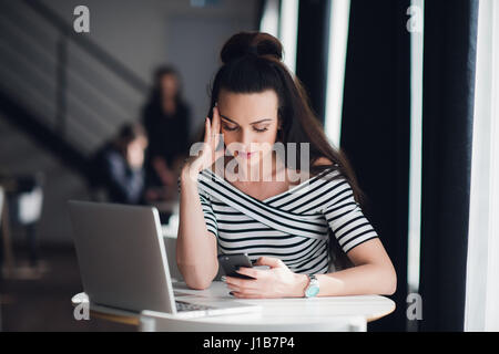 Schöne junge Geschäftsfrau mit Handy während der Arbeit mit Laptop im Büro, sie ist konzentriert, versucht, das Problem zu lösen. Verärgert oder enttäuscht Ausdruck auf ihrem Gesicht. Moderne Büro-Landschaft. Stockfoto