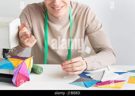 Quilten und Handarbeiten - close-up auf den Händen der lächelnden männliche Schneider, seine Finger Faden in die Nadel hängen um den Hals, Maßband, auf Tabelle Stücke genäht, Stoffe und Spulen von threads Stockfoto