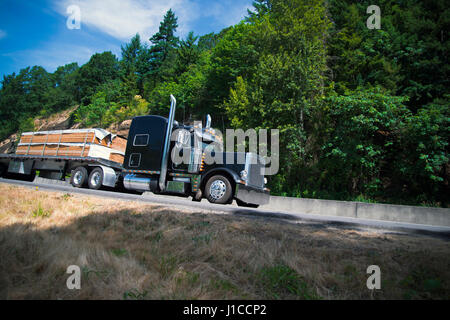 Schwarze elegante Sattelschlepper mit Chrom Zubehör individuelle Gestaltung transportiert die Holz Balken auf dem Tieflader auf der schönen Autobahn bewegen Stockfoto