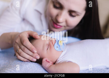 Neugeborenes Baby junge mit Mutter. Mama sanft streichelte den Kopf des Babys. Baby süß schlafend auf einem weißen Bett. Kind mit einem Schnuller im Mund. Stockfoto