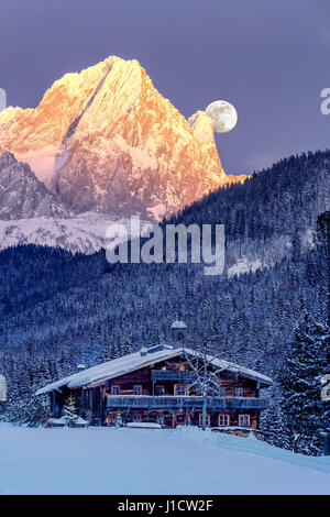 Vollmond steigt hinter den Schnee-caped Alpen mit alten, traditionellen Bauernhaus im Vordergrund. Stockfoto