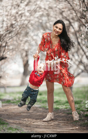 Junge Mutter mit ihrem Baby auf Spaziergang im blühenden Garten. Sie spielen und Lachen fröhlich unter Blütenbäumen Aprikose. Stockfoto