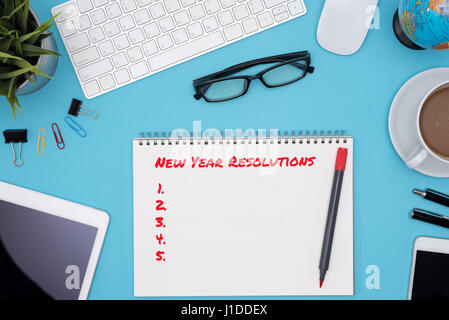 Neujahr Resolutionen Konzept mit offen Spiral-Notizbuch, Computer, Stift, Handy, Tablet, Blume, Brille, Globus und Kaffee Augenmuschel auf blauer Schreibtisch ta Stockfoto