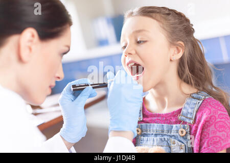 Frau Doktor untersucht Hals des kleinen Mädchens mit Spachtel und Taschenlampe Stockfoto