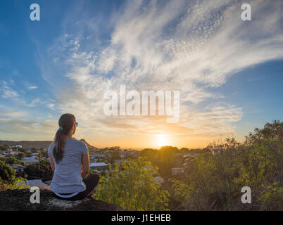 Eine junge Frau sitzt auf einem Felsvorsprung, einen wunderschönen Sonnenuntergang über Honolulu, Hawaii, mit Diamond Head im Hintergrund teilweise sichtbar. Stockfoto