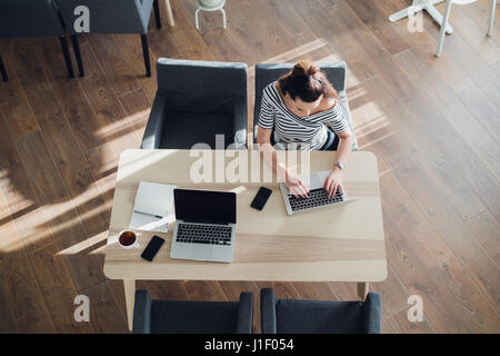 Direkt oberhalb der Ansicht der weiblichen Eingabe auf Laptop. Laptop, digital-Tablette, Tagebuch, Kaffeetasse und zwei Smartphones am Schreibtisch. Woan von zu Hause aus arbeiten. Stockfoto