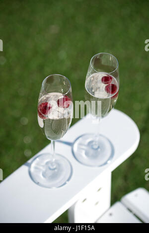 Zwei Gläser Prosecco mit Himbeeren auf weißen Gartenstuhl mit grünen Rasen. Stockfoto