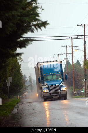 Große moderne blau semi-LKW mit Anhänger und Scheinwerfer und Reflexion auf nasser Fahrbahn mit Holz-Stangen von Stromleitungen und Bäumen und grauen Himmel Stockfoto