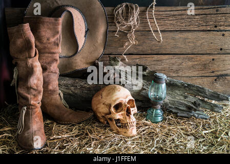 Stillleben mit menschlichen Schädel auf Heu mit traditionellen Lederstiefel und Westamerika Rodeo braune Cowboyhut Hintergrund, Vintage und dunklen Ton für horro Stockfoto