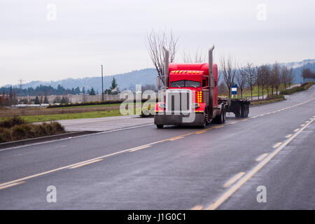 Stilvolle rote klassische big Rig Sattelschlepper mit vertikalen Auspuffrohre und die Reihen von orange Seitenscheiben und einem Anhänger auf der Straße mit einer Aufteilung Streifen Stockfoto