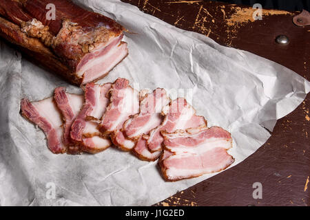 Ansicht-Scheiben geräucherter Speck auf dem weißen Verpackungspapier hautnah. Naturmort im rustikalen Stil auf braune Vintage-Hintergrund. Stockfoto