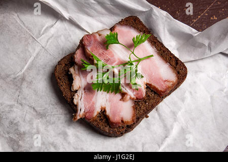 Nahaufnahme Blick Scheiben geräucherter Speck am Stück Roggenbrot mit Kräutern - rustikalen Stil auf braune Vintage-Hintergrund. Stockfoto