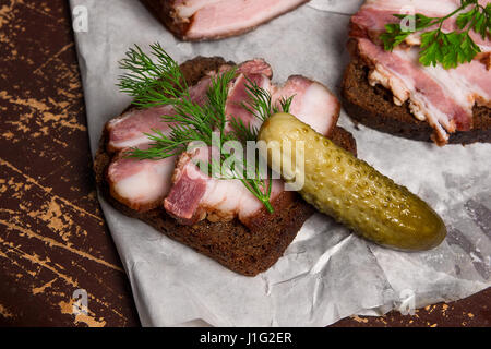 Nahaufnahme Blick Scheiben geräucherter Speck am Stück Roggenbrot mit Kräutern und mehrere Gurken - rustikalen Stil auf braune Vintage-Hintergrund. Stockfoto