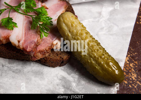 Nahaufnahme Blick Scheiben geräucherter Speck auf das Stück Roggenbrot mit Kräutern, mehrere Gurken, altes Messer mit Holzgriff - rustikalen Stil auf braun vintage Stockfoto